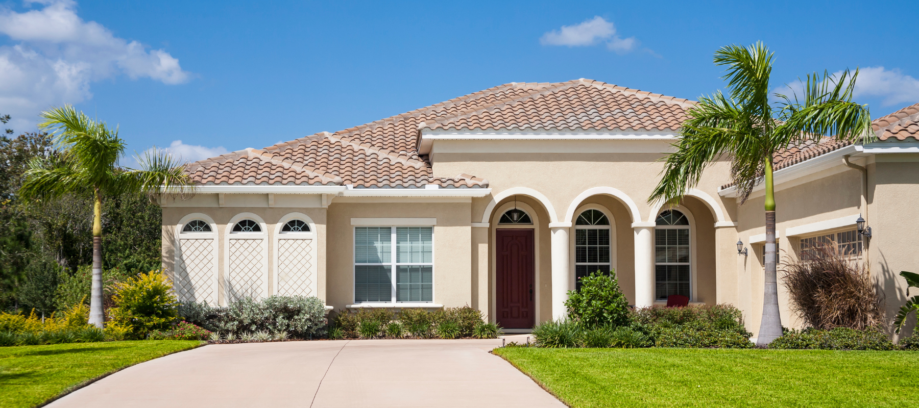A beige stucco home with a terracotta tile roof, framed by palm trees and vibrant landscaping