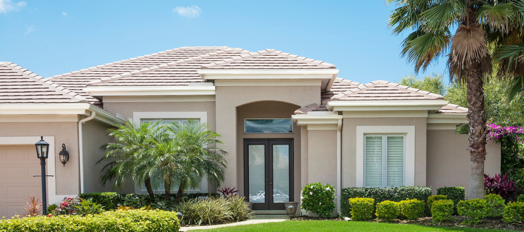 A taupe stucco home with a tile roof and manicured shrubs, featuring a welcoming double-door entrance