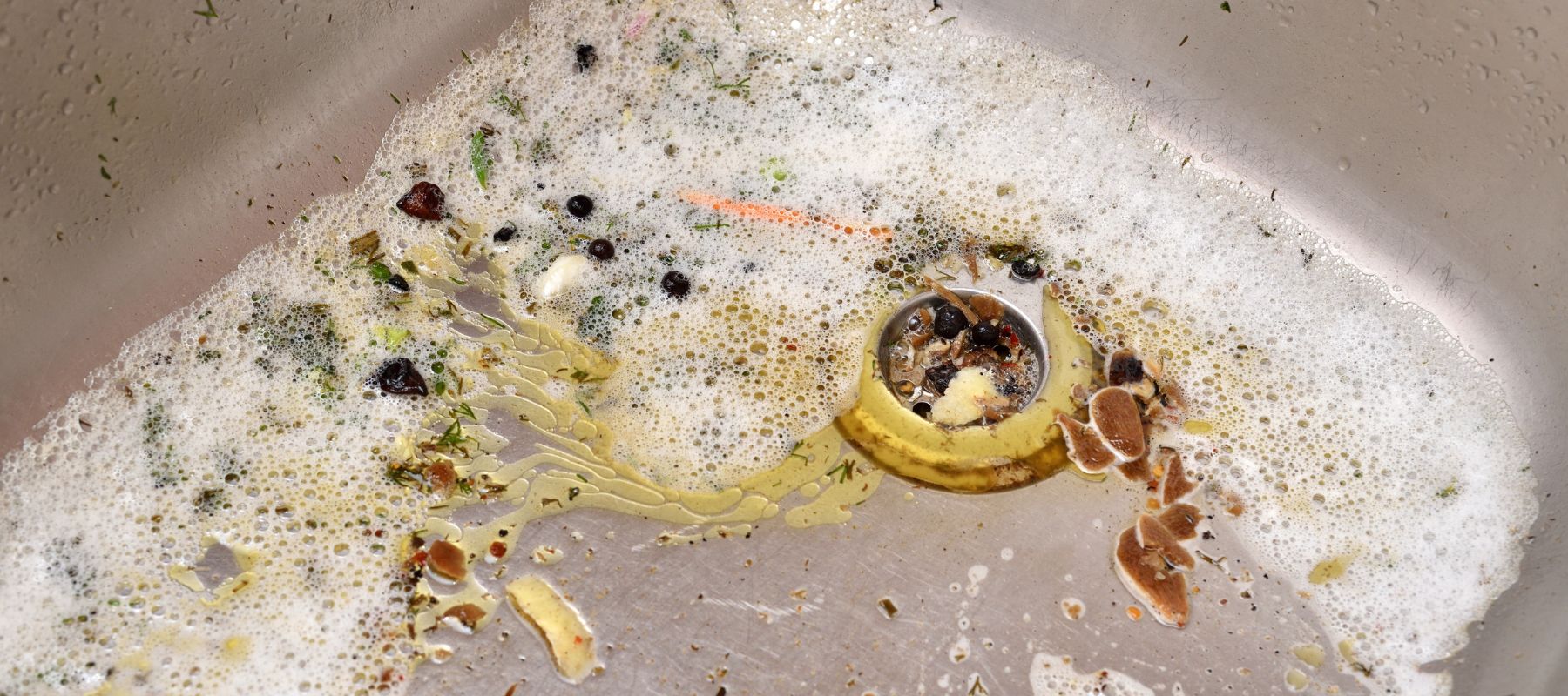 Close-up of a kitchen sink drain filled with food scraps, grease, and soapy water, showing a common cause of clogs