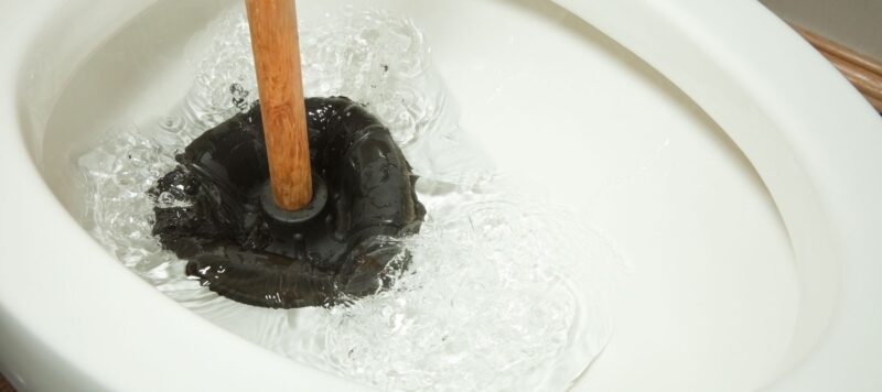 A toilet being plunged with a black rubber plunger in swirling water to clear a clog