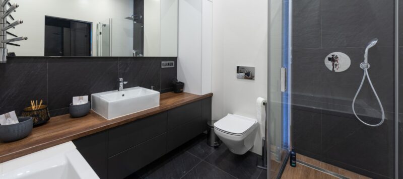 A sleek modern bathroom features a dark vanity with a white rectangular sink, a wall-mounted toilet, and a glass-enclosed shower with dark gray tiles